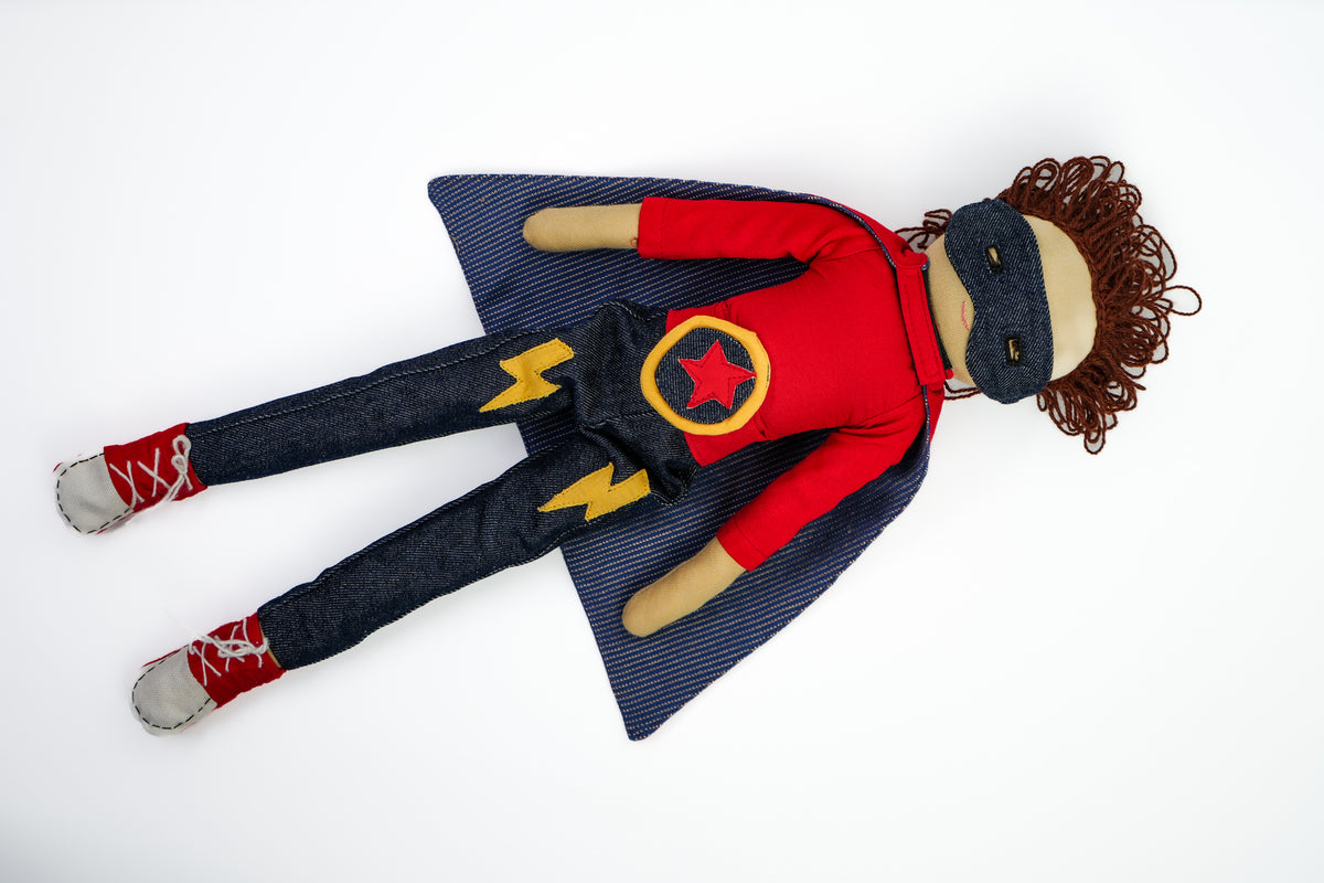 A handmade felt doll dressed as a superhero, featuring a red long-sleeve shirt with a yellow star emblem, dark blue pinstriped cape, dark blue jeans, red sneakers, and a black mask, laid flat on a white background.