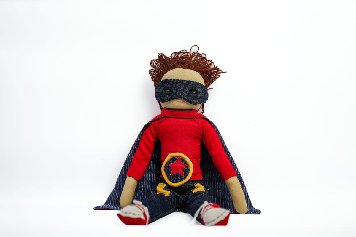 A handmade fabric superhero doll with curly brown hair, wearing a red shirt, dark blue cape and mask, and boots, photographed against a plain white background.