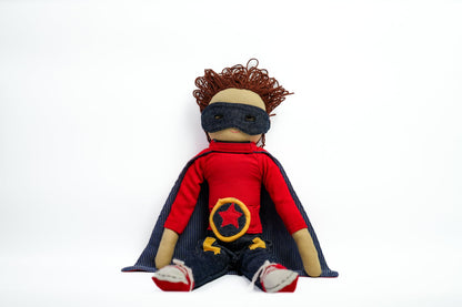 A handmade fabric superhero doll with curly brown hair, wearing a red shirt, dark blue cape and mask, and boots, photographed against a plain white background.