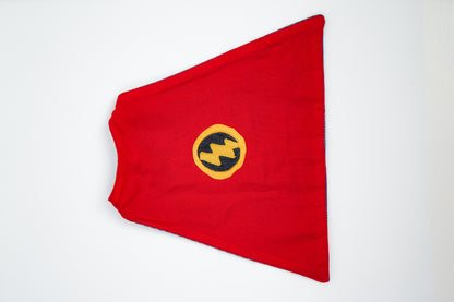 A red fabric cape laid flat on a white background, featuring a circular yellow and black emblem with a lightning bolt motif in the center.